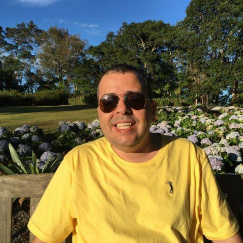 Fotografia colorida em formato circular de Décio Nascimento Guimarães, sentado em um banco de praça é visto da cintura para cima, atrás dele, flores e árvores. Ele é branco, tem cabelos pretos e curtos, nariz médio e sorri. Usa óculos escuro e camiseta amarela.